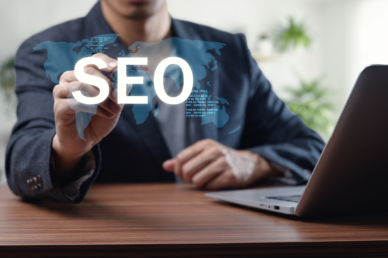 A businessman interacts with a digital SEO concept displayed on a virtual screen while seated at an office desk with a laptop, illustrating the significance of search engine optimization in modern marketing strategies.
